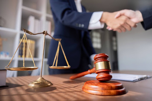 A man shakes the hand of another over a legal desk that has a gavel and the scales of justice.